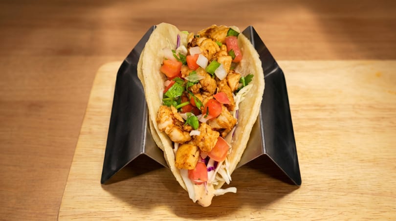 A chicken taco with diced tomatoes, onions, and cilantro in a metal holder on a wooden surface.