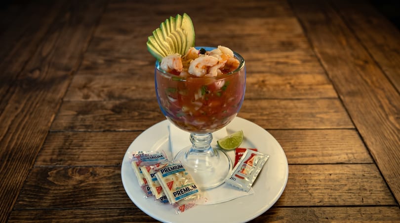 Shrimp cocktail in a glass with avocado, lime, and cracker packets on a white plate.