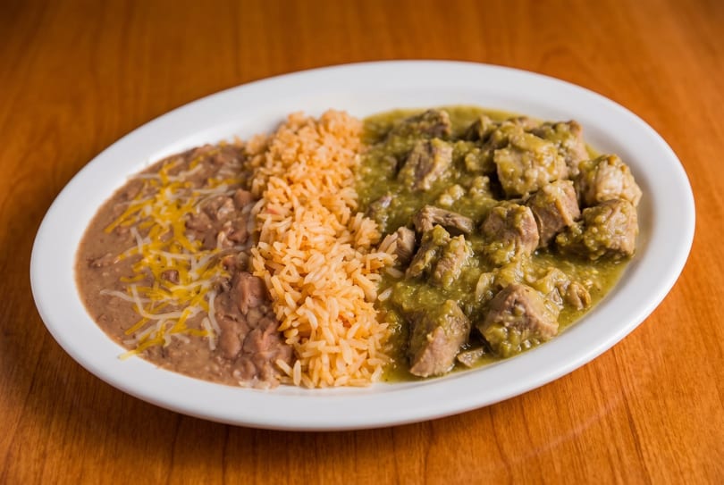 A white oval plate with chile verde, Spanish rice, and refried beans topped with cheese.
