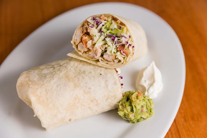Two chicken burritos, one cut open, with guacamole and sour cream on a white plate.
