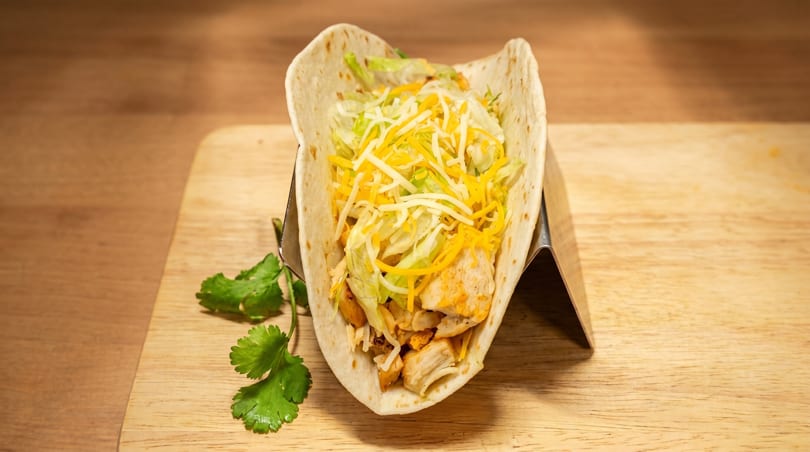 A chicken taco with shredded lettuce and cheese in a soft tortilla, on a wooden board.
