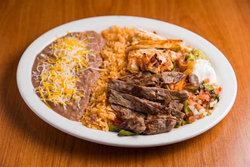 A white plate with steak, chicken, rice, refried beans with cheese, guacamole, and pico de gallo.