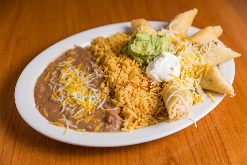A white plate with refried beans, yellow rice, guacamole, sour cream, and taquitos.