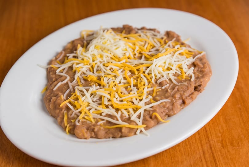 Refried beans topped with shredded yellow and white cheese on a white oval plate.