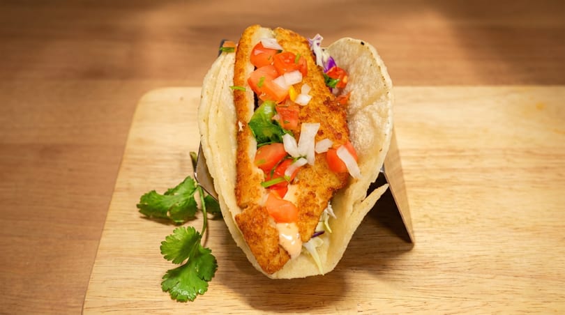 Crispy fish taco with diced tomatoes, onions, and cilantro, served on a wooden board.