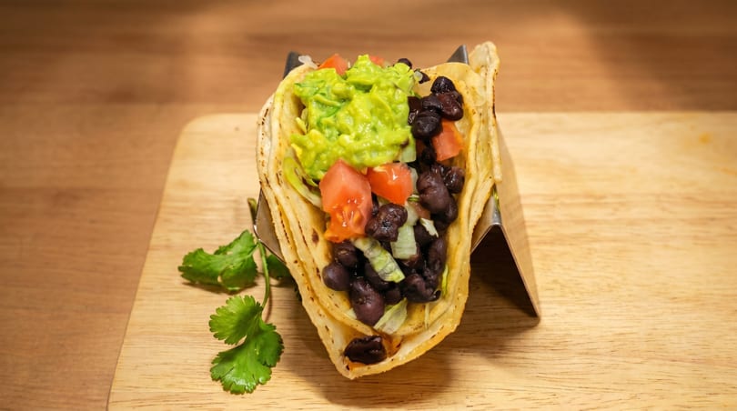 A vibrant taco filled with guacamole, black beans, diced tomatoes, and lettuce on a wooden board.